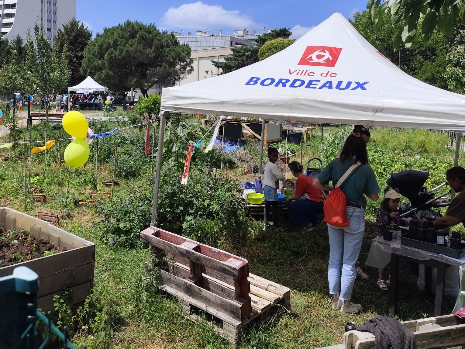 Un barnum au milieu du jardin participatif avec un banc en palettes, des familles visitent